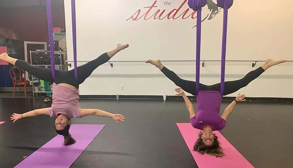 Two people are performing aerial yoga poses using purple silk hammocks in a studio with one person inverted and the other balancing on her head