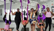 A group of smiling people are using purple aerial silks in a fitness studio, with one person doing an upside-down pose.