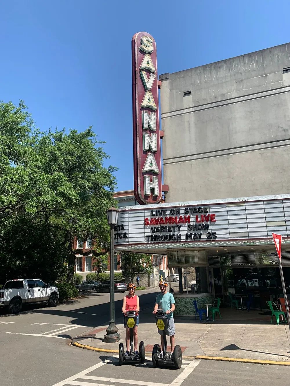 Two people are riding Segways in front of the historic Savannah Theater with a luminescent sign under a clear blue sky