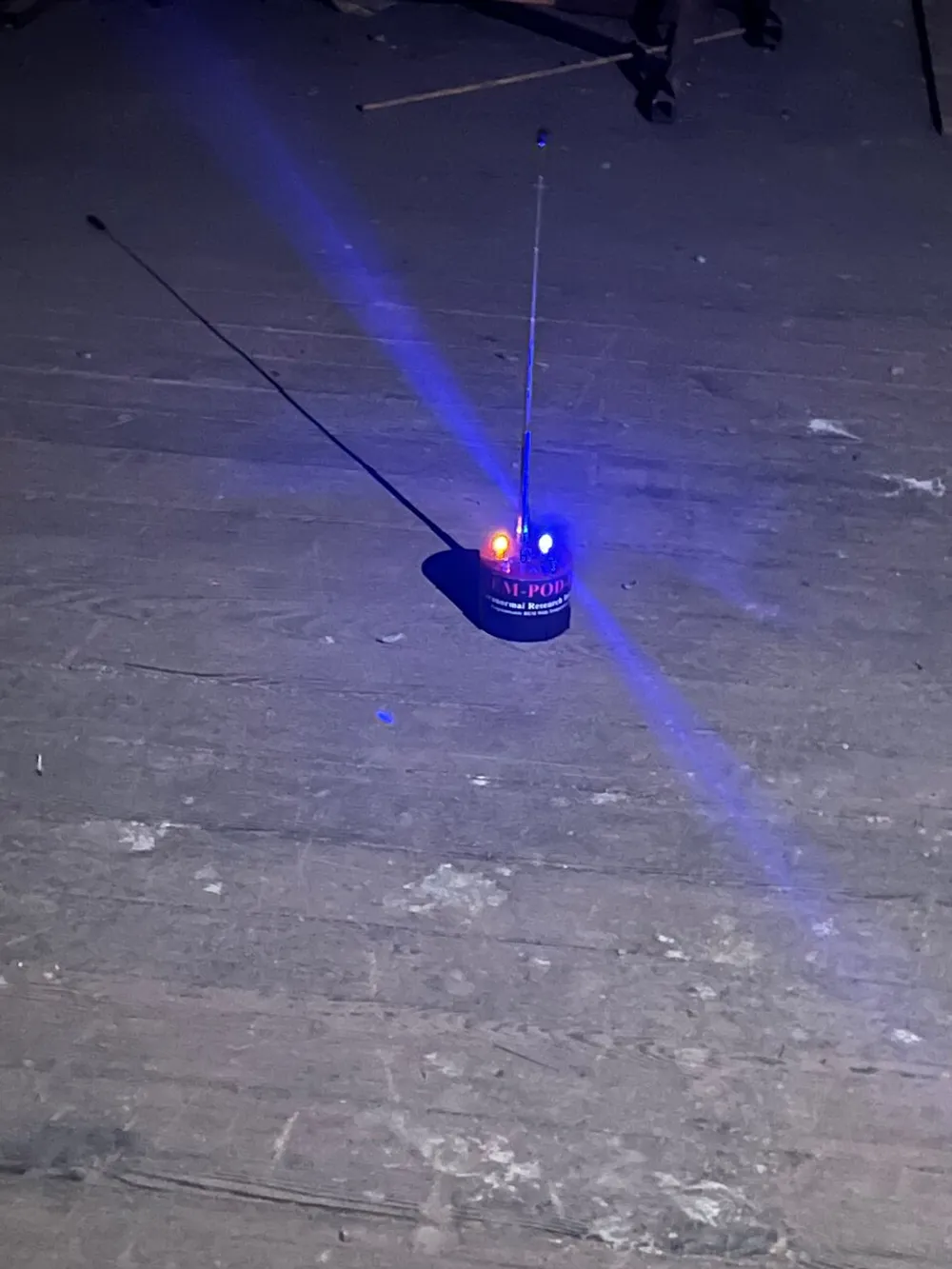 This image shows a small robotic device with flashing red and blue lights on a wooden floor projecting a blue light beam