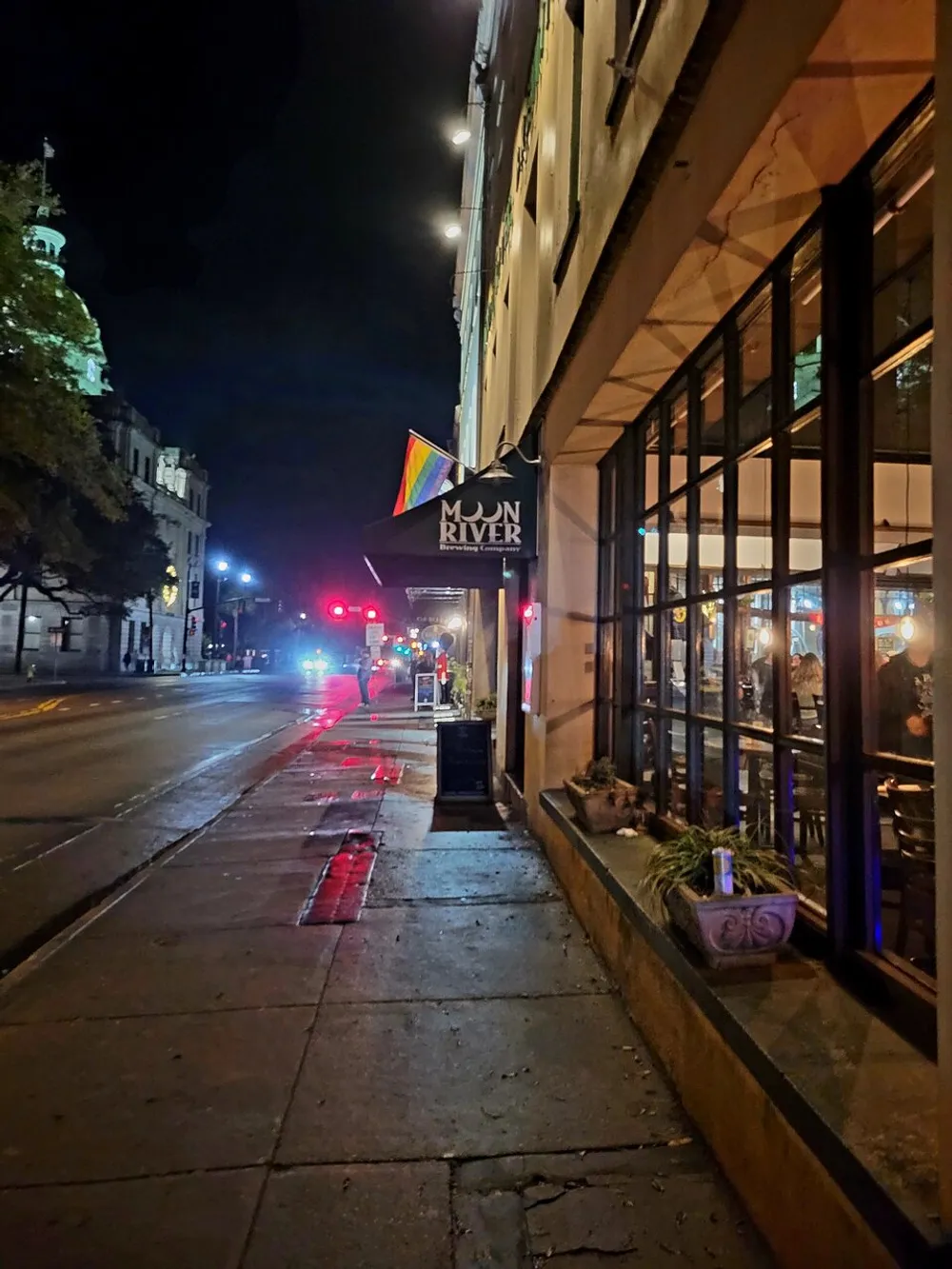 This image shows a city street at night with the exterior of the Moon River Brewing Company and a pride flag hanging above alongside illuminated windows and a view of the street receding into the distance with traffic lights and a glimpse of a historic buildings tower