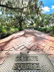 The image shows a sunny pathway lined with brick pavers leading through a park with dangling Spanish moss from large oak trees, with a metal plate in the foreground that reads 
