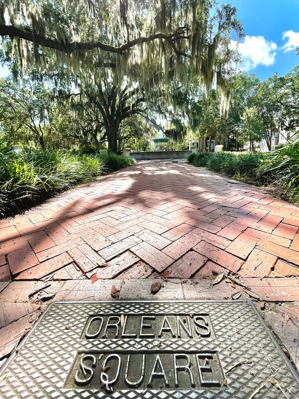 The image shows a sunny pathway lined with brick pavers leading through a park with dangling Spanish moss from large oak trees with a metal plate in the foreground that reads Orleans Square