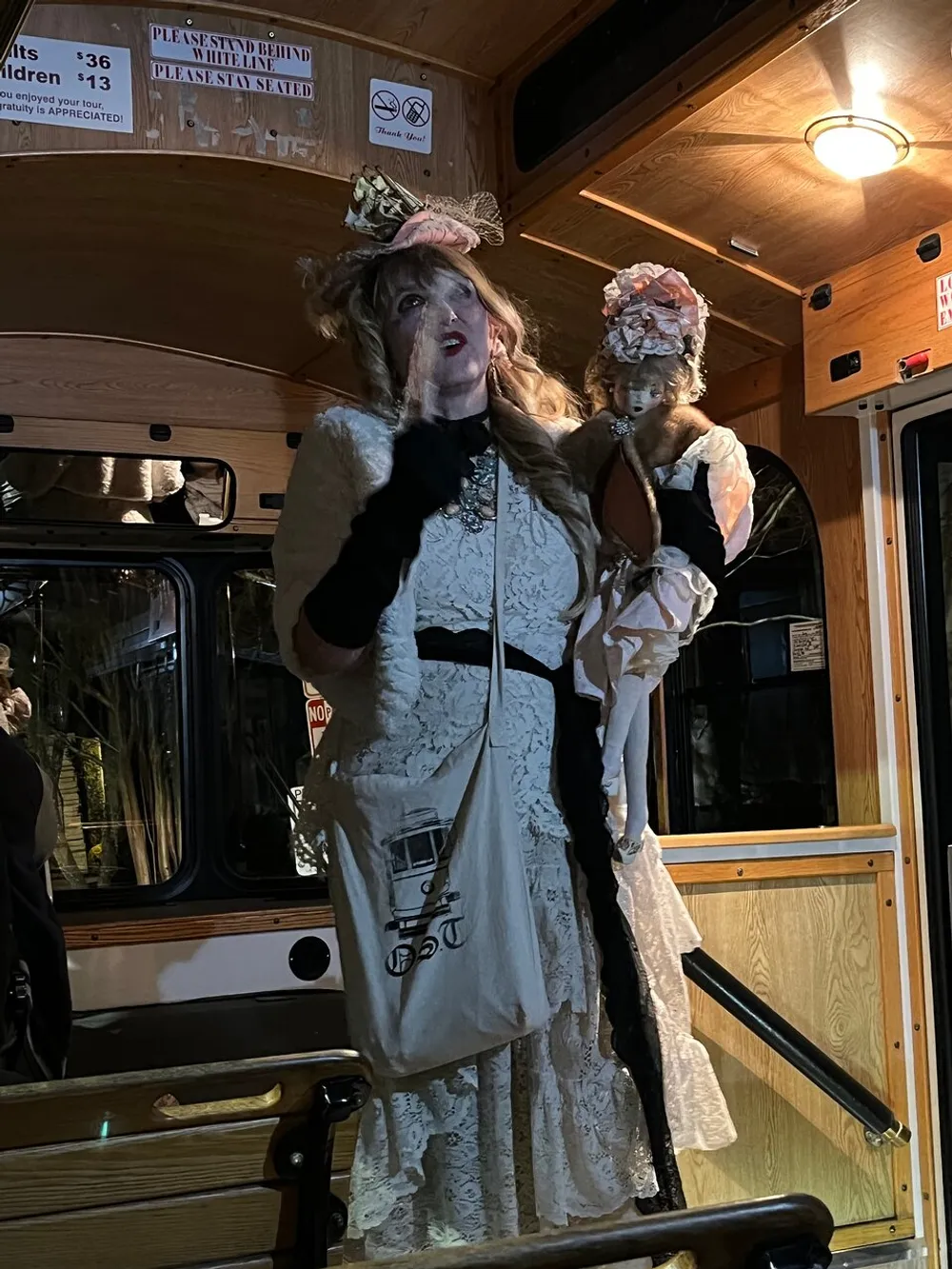 A person dressed in vintage attire complete with a hat and holding a doll is standing on a vehicle resembling a trolley or bus surrounded by warm lighting and wooden paneling