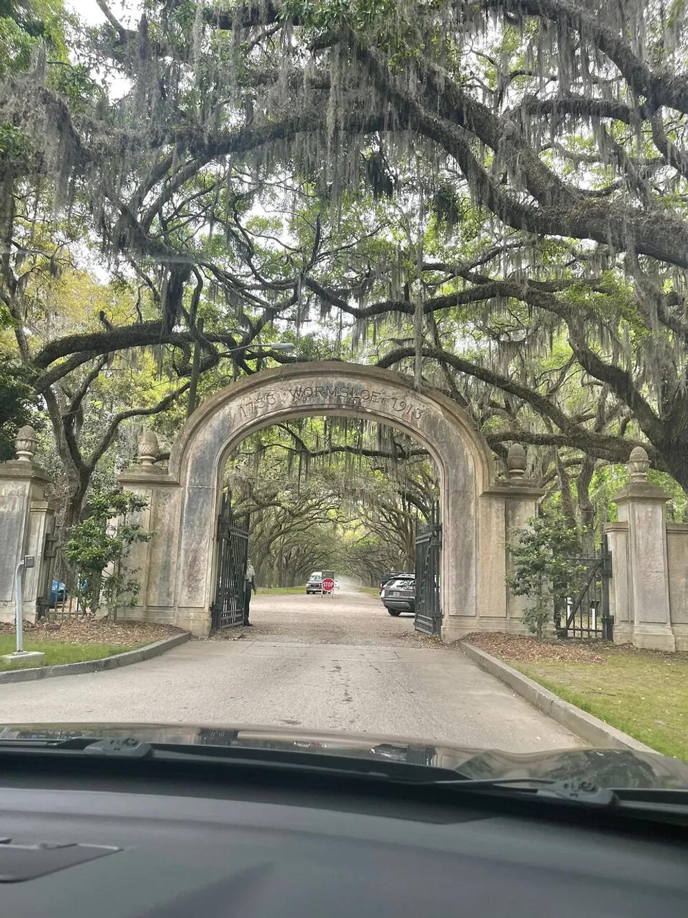 The image shows a view from a car approaching an ornate stone gateway labeled Wormsloe 1733 flanked by a canopy of trees draped with Spanish moss