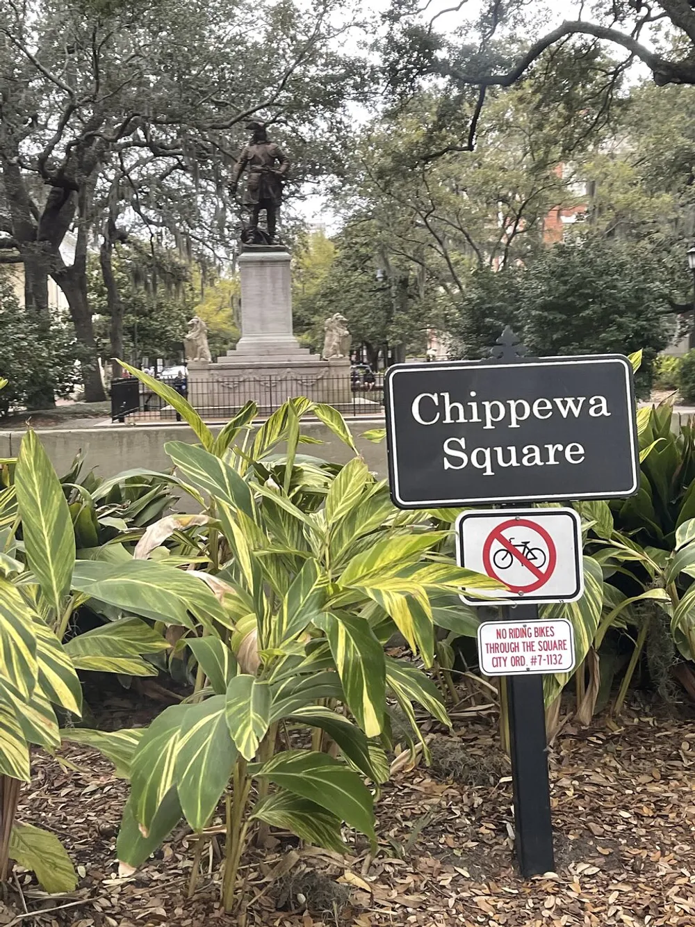The image shows a sign for Chippewa Square with lush greenery in the foreground and a statue of a historical figure on a pedestal in the background surrounded by the ambiance of a peaceful park