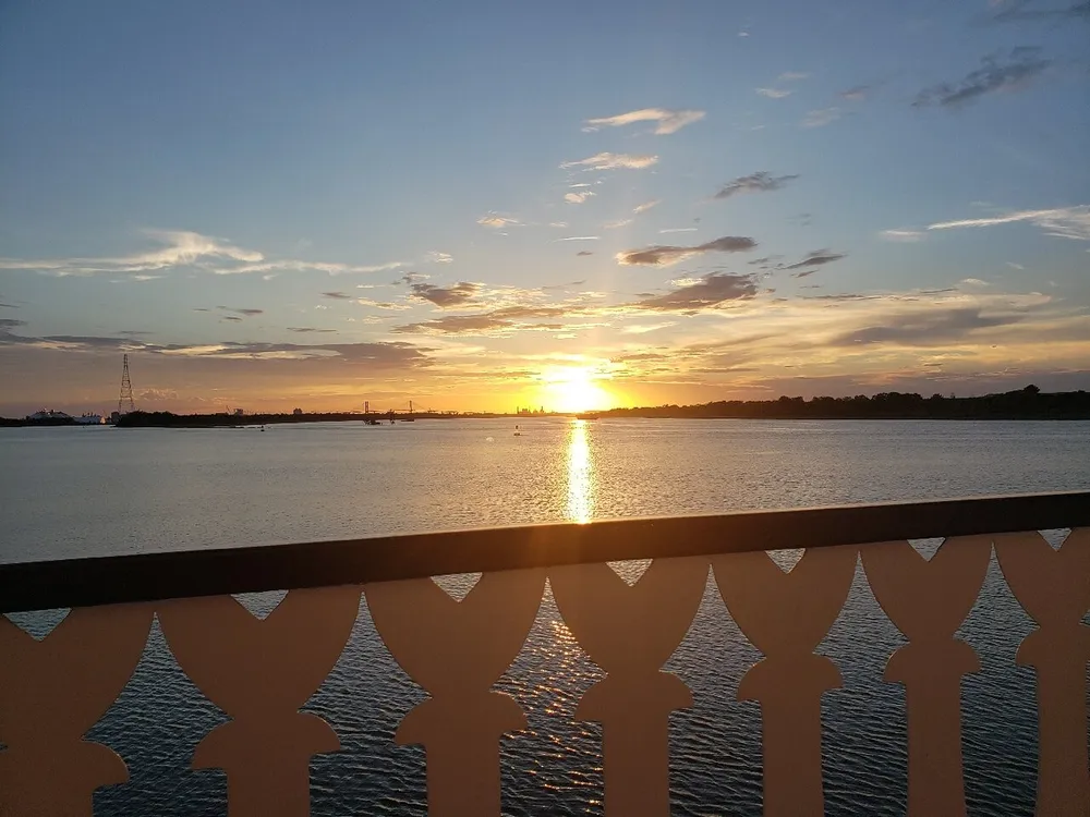 The image captures a tranquil sunset over a calm body of water viewed through an ornamental railing with clouds scattered across the sky and the sun reflecting on the water