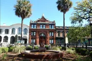 The image features a picturesque urban square with a lion sculpture fountain in the foreground and a historic red-brick building flanked by palm trees under a clear blue sky.