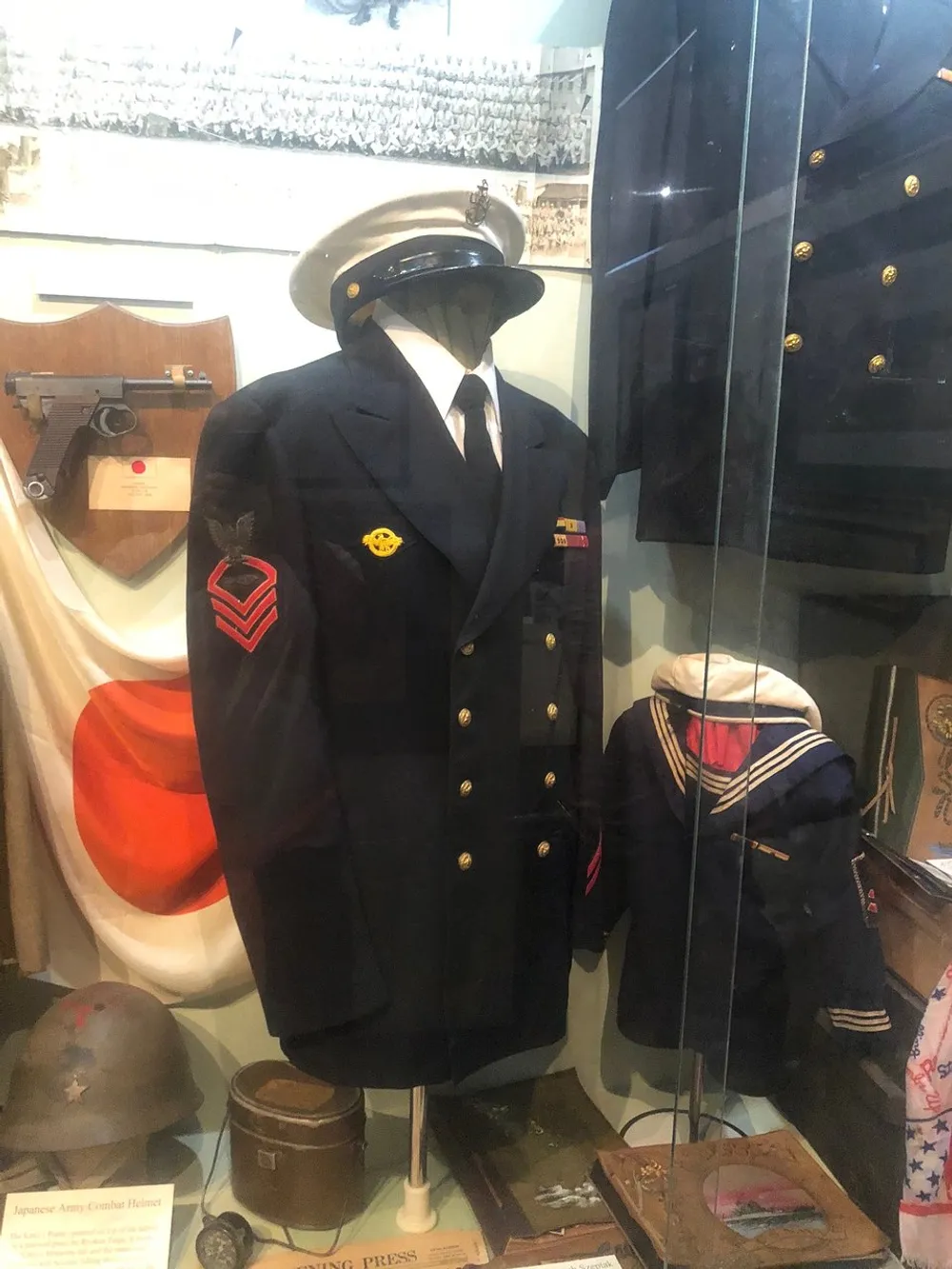 The image shows a collection of military uniforms and artifacts displayed in a glass case with a prominent navy jacket and hat on a mannequin suggesting a historical or educational exhibit possibly within a museum