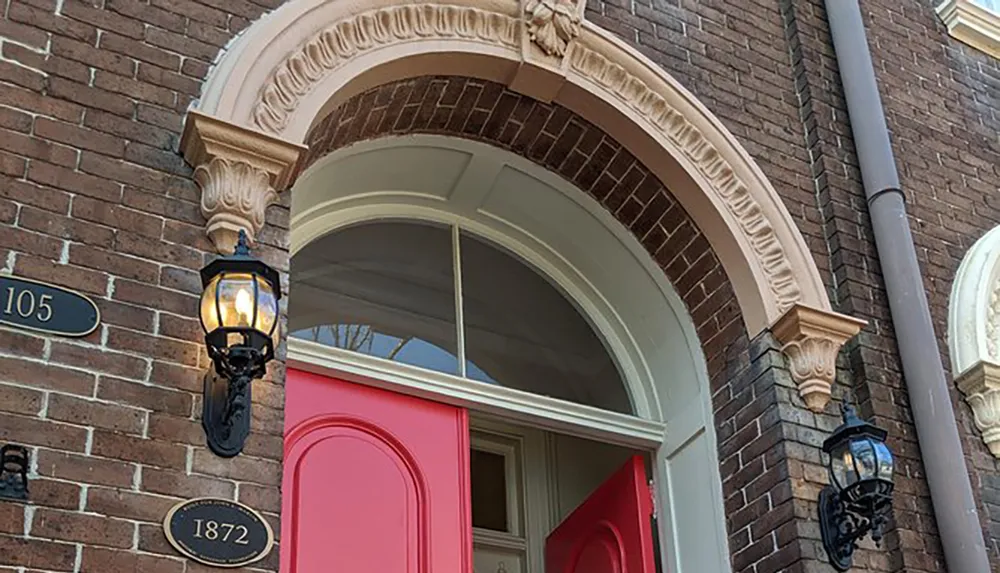 The image shows a red door on a brick building with a transom window above it flanked by two classic lantern-style wall sconces with plaques displaying 105 and 1872 near the entryway