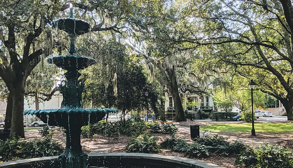 The image shows a tranquil park with a tiered fountain in the foreground dappled sunlight filtering through the lush canopy of Spanish moss-draped trees with a glimpse of urban life in the background
