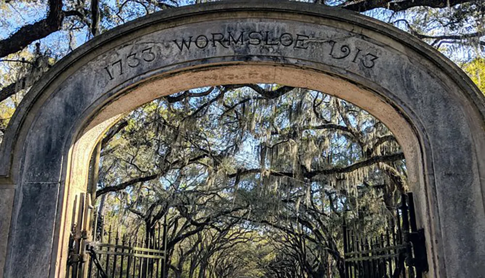 The image shows an ornate stone arch inscribed with 1733 WORMSLOE 1913 framing a view of a tree-lined path draped with Spanish moss