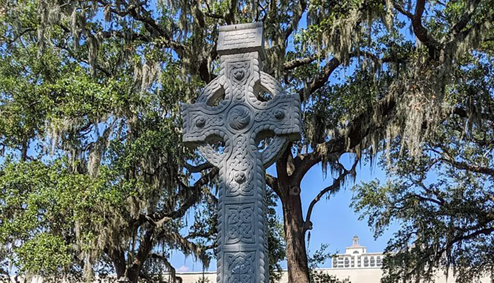An intricately carved stone Celtic cross stands tall amidst Spanish moss-draped trees under a clear blue sky