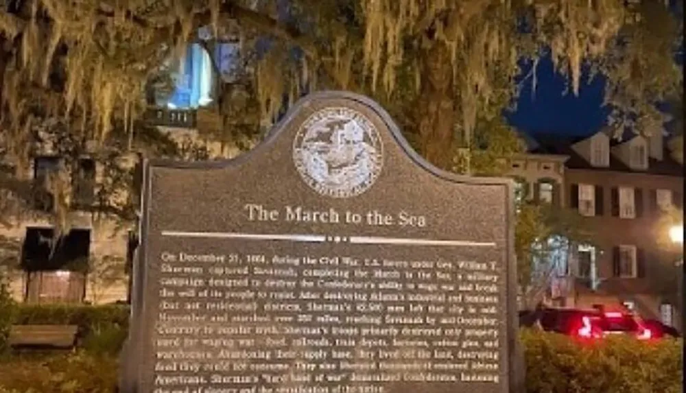 The image shows an informational plaque titled The March to the Sea commemorating a significant event from the American Civil War with hanging Spanish moss and a historic building illuminated in the background