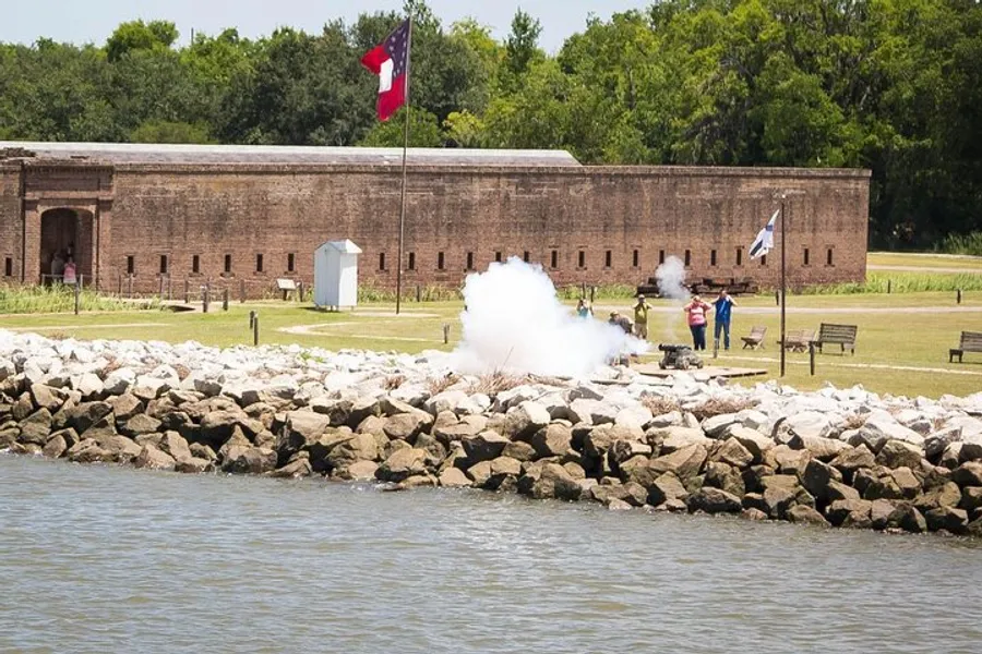 This image shows a historic fort with a large Texas flag flying, people nearby, and a cannon firing, creating a large cloud of smoke along the rocky shoreline.