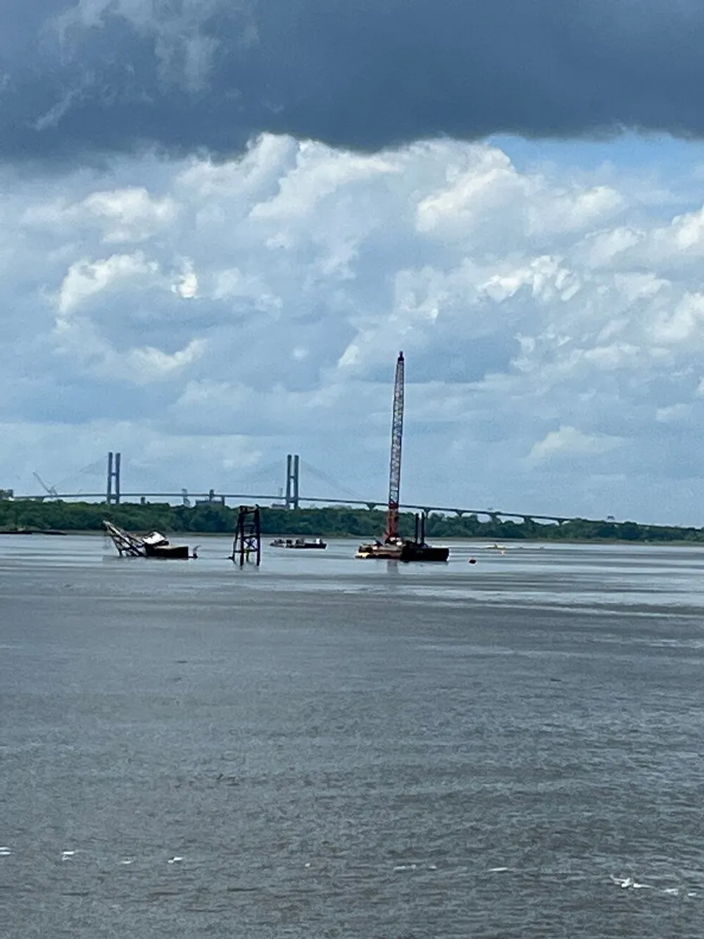 The image shows a body of water with machinery and a structure resembling a crane under a sky with a mix of clouds and blue patches and a faintly visible bridge in the distant background