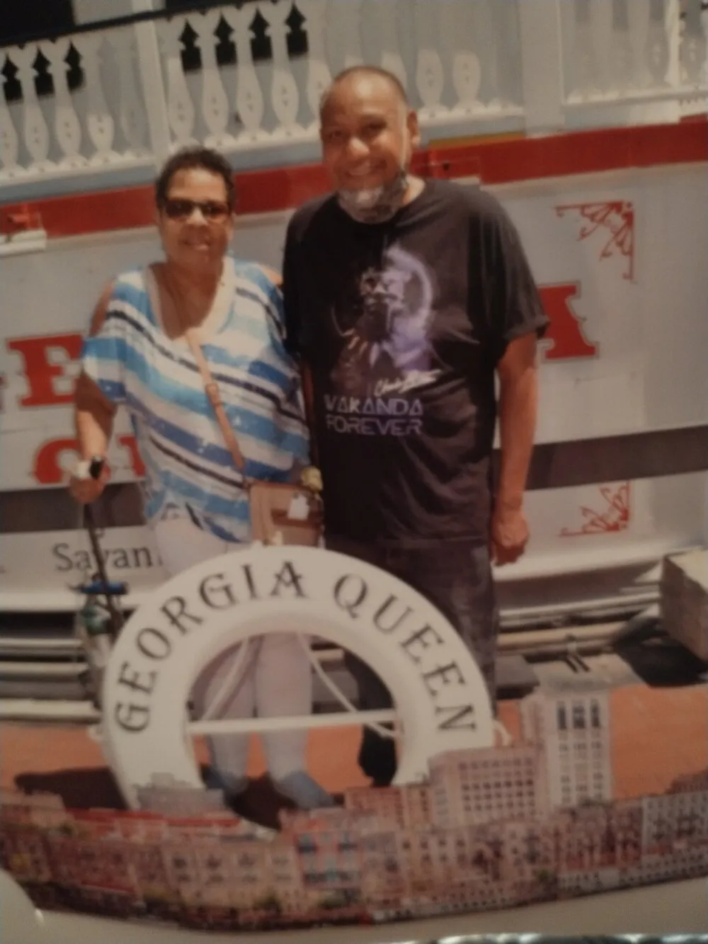 A smiling man wearing a Wakanda Forever t-shirt and a woman in a striped top are posing in front of a lifebuoy labeled GEORGIA QUEEN likely taken on a riverboat or at a waterfront attraction