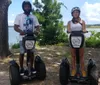 A man and a woman pose for a photo while standing on their respective Segways in a park setting with trees and a body of water in the background