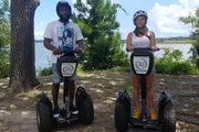 A man and a woman pose for a photo while standing on their respective Segways in a park setting with trees and a body of water in the background.