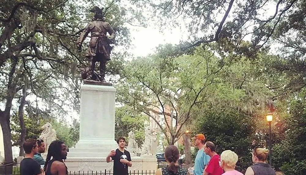 A group of people is gathered for a tour or presentation in a park in front of a statue with verdant trees surrounding them