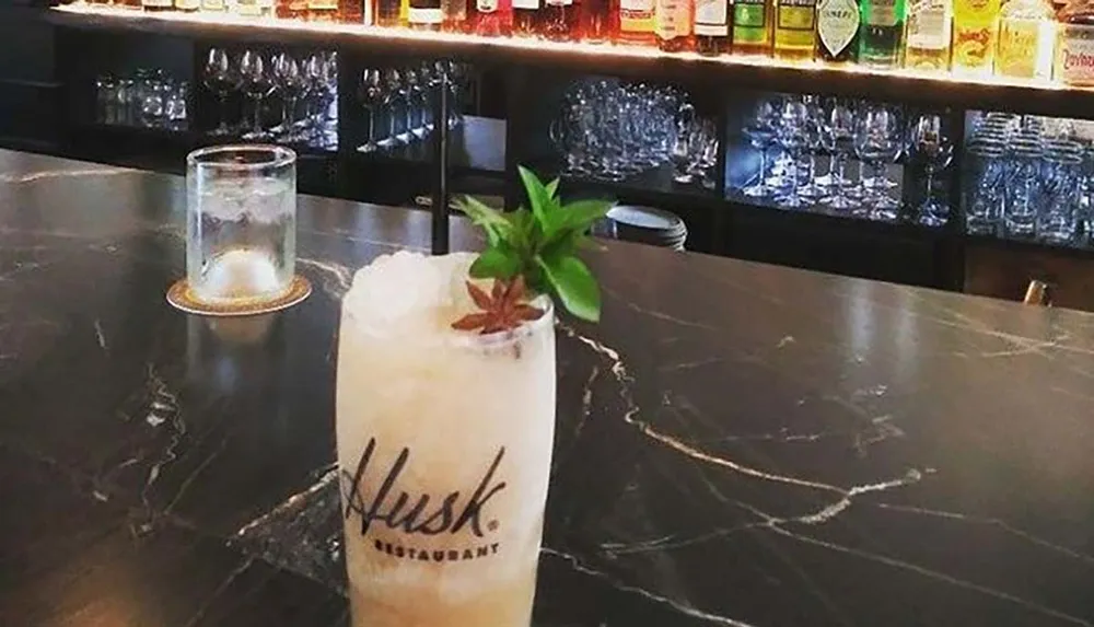 The image shows a cocktail with a sprig of mint on a bar counter with a backdrop of shelves stocked with various bottles of liquor and glassware