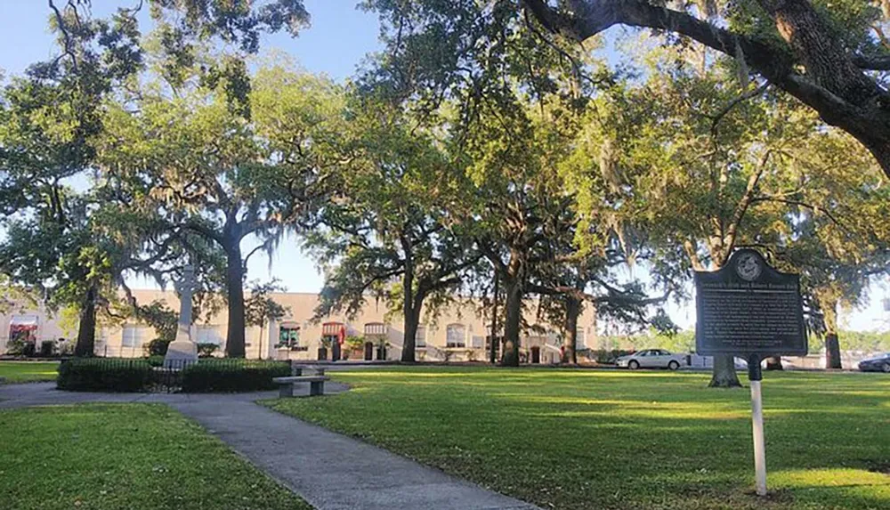 The image shows a tranquil park with lush green grass large oak trees draped with Spanish moss a few benches and a historical marker in the foreground