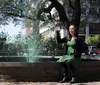 A woman in a green dress and black jacket is smiling and sitting on a fountains edge seemingly toasting with a glass with green-tinted water spouting behind her likely in celebration of St Patricks Day