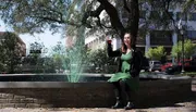 A woman in a green dress and black jacket is smiling and sitting on a fountain's edge, seemingly toasting with a glass, with green-tinted water spouting behind her, likely in celebration of St. Patrick's Day.