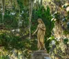 A weathered statue of a woman stands gracefully among verdant trees draped with Spanish moss evoking a serene and timeless atmosphere