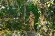 A weathered statue of a woman stands gracefully among verdant trees draped with Spanish moss, evoking a serene and timeless atmosphere.