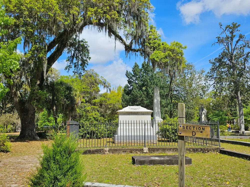 The image depicts a tranquil cemetery scene marked as Section D with a mausoleum lush greenery and Spanish moss-draped oak trees under a clear blue sky
