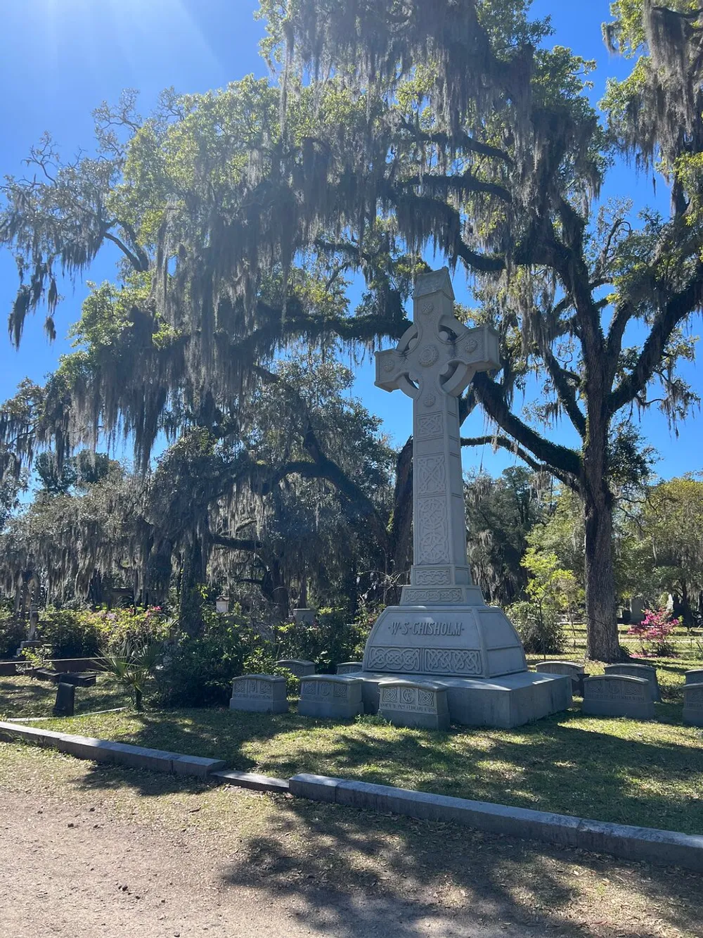 The image shows a tall ornate cross gravestone in a cemetery with Spanish moss-draped trees in the background under a clear blue sky