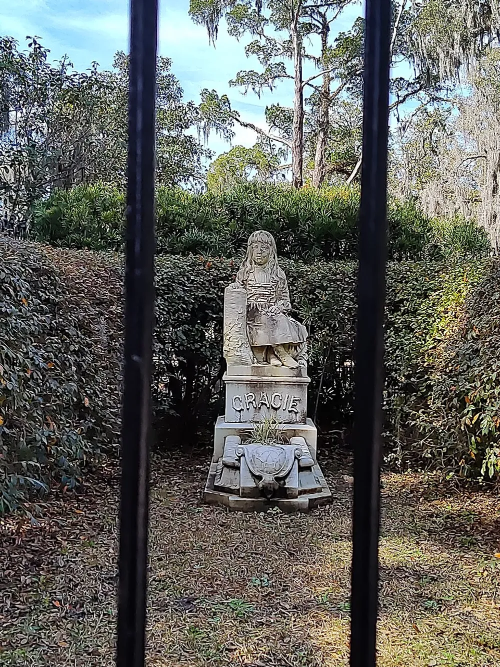 The image shows a sculpture of a young girl sitting on a bench labeled Gracie framed by vertical bars in the foreground and surrounded by trees and shrubbery