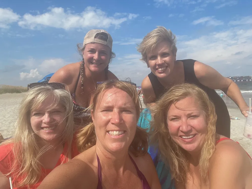 Five smiling individuals are taking a group selfie on a sunny beach with the ocean in the background