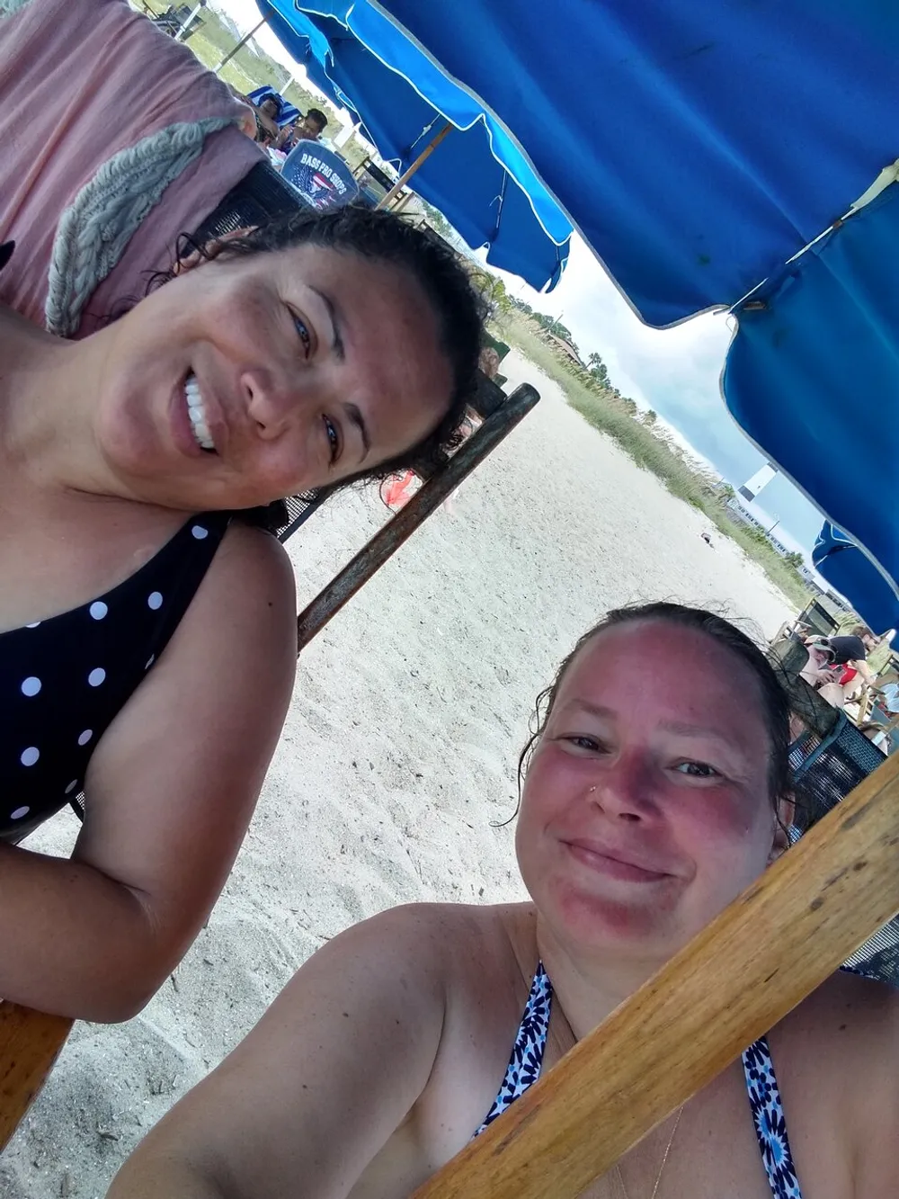 Two smiling individuals are taking a selfie under blue beach umbrellas on a sandy shore