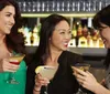 Three women are smiling and toasting with cocktails at a bar