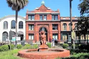 An ornate red brick building with a fountain featuring a lion statue in front is situated in a sunny square with palm trees and pedestrians walking by.