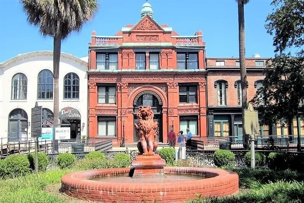 An ornate red brick building with a fountain featuring a lion statue in front is situated in a sunny square with palm trees and pedestrians walking by