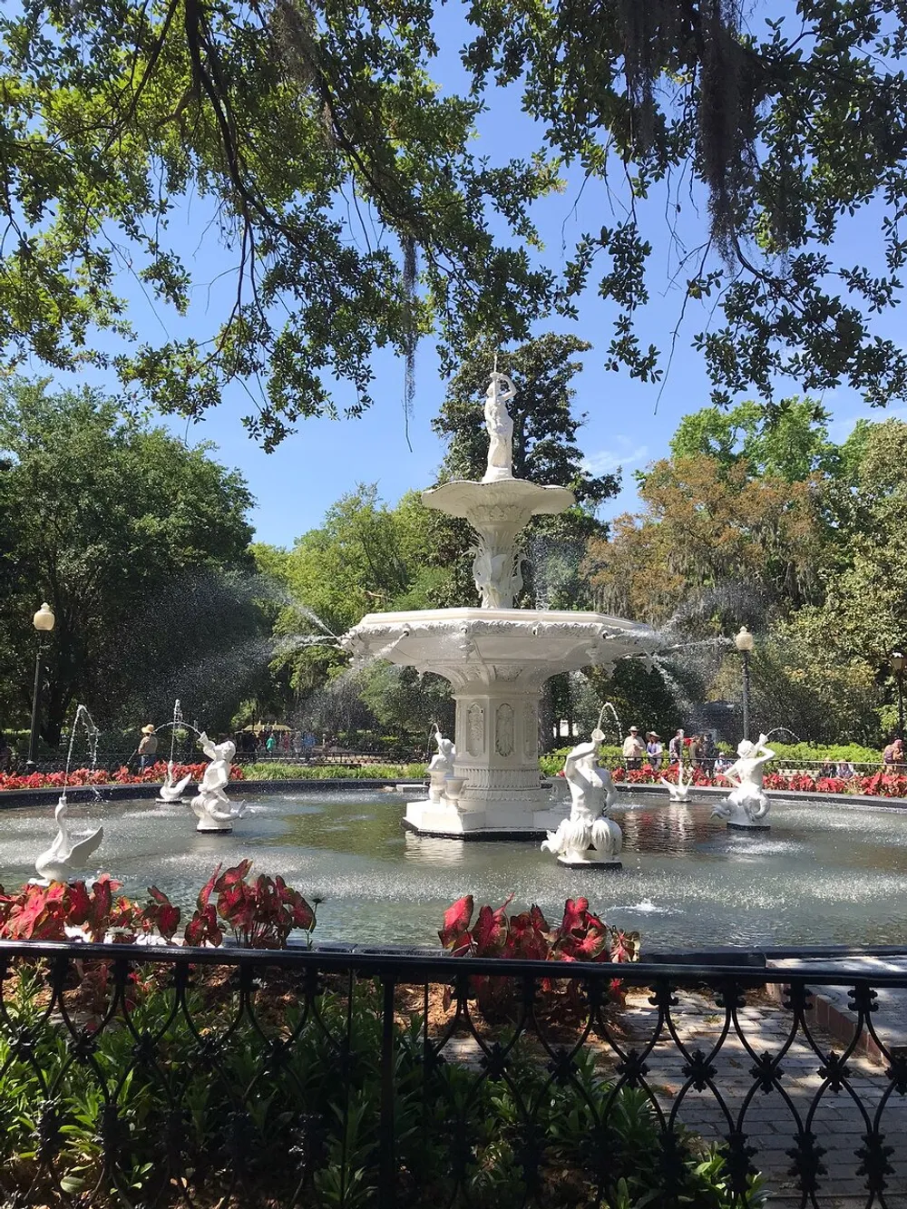 The image shows an ornate white fountain with water jets surrounded by greenery and red flowers framed by tree branches above and a black fence in the foreground on a sunny day