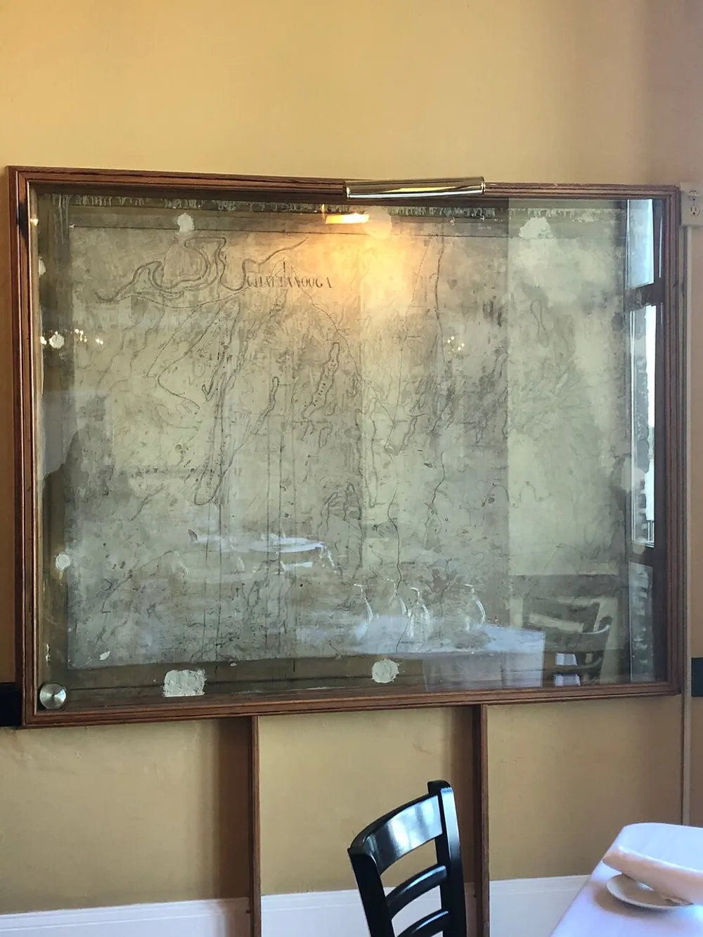 This image shows a framed worn and aged document or drawing mounted on a wall viewed through a reflective glass surface which reveals some items in the room