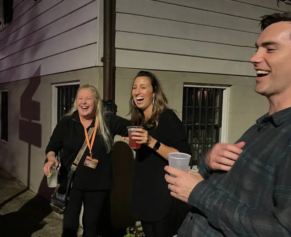 Three people are laughing and enjoying a social gathering at night holding drinks in their hands