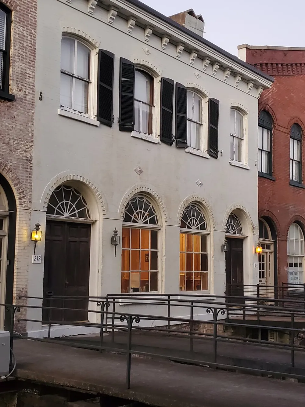 The image shows a two-story building with arched windows and doors black shutters decorative brickwork and a ramp leading to the entrance all suggesting historical architecture captured in the dimming light of dusk