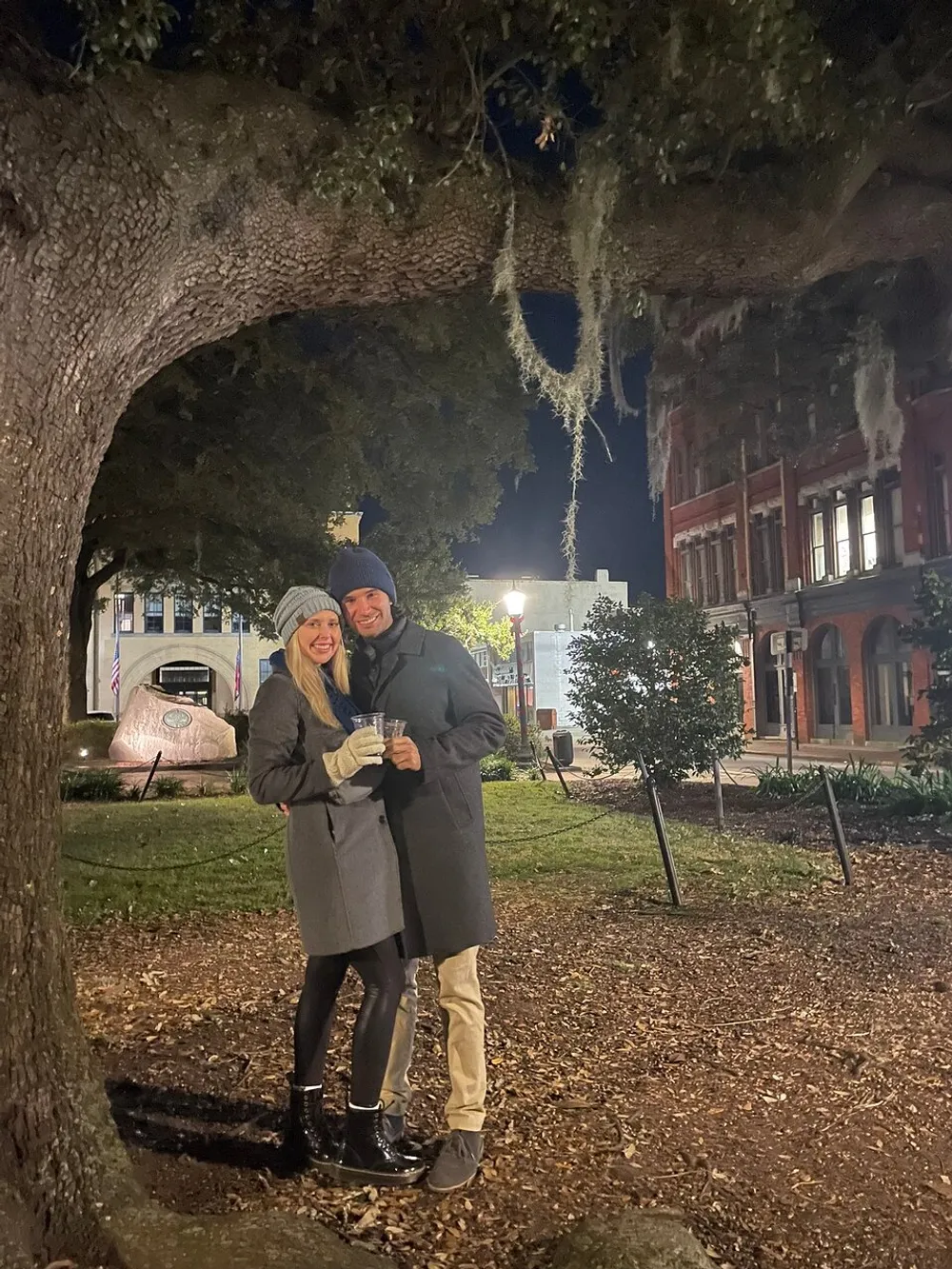 A smiling couple stands closely together under a tree adorned with Spanish moss at night evoking a cozy and romantic atmosphere