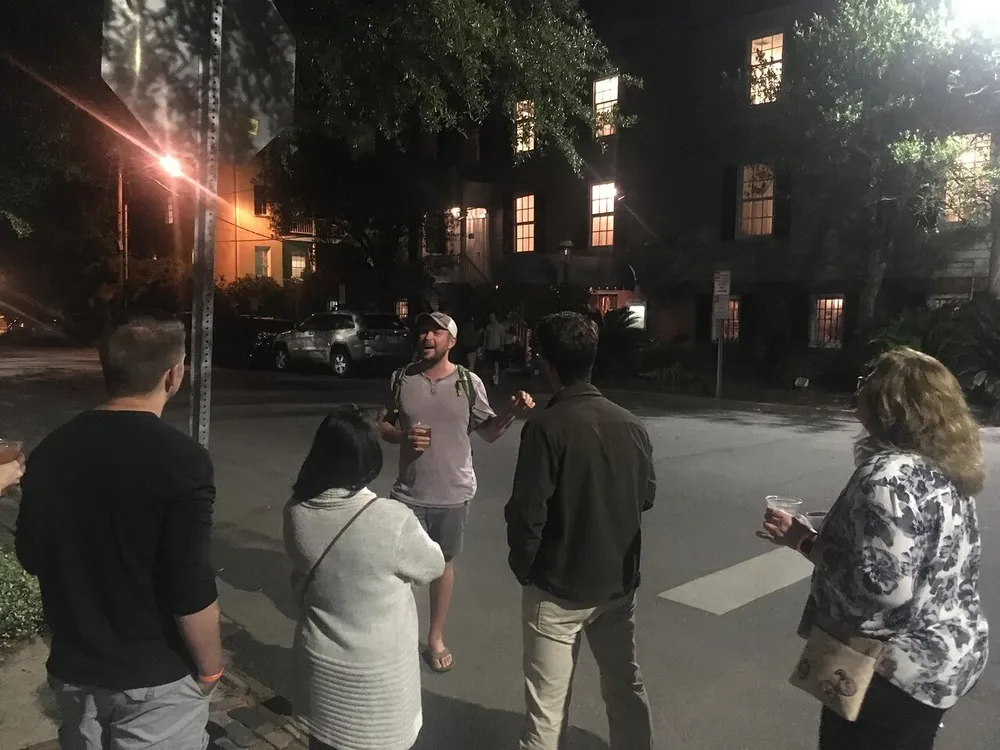 A group of people appear to be on a night tour or gathering with one person speaking to the others while they listen attentively on a street lit by streetlights and illuminated windows