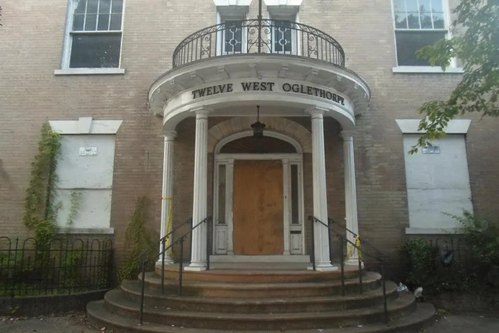 The image shows the entrance of a brick building with a curved portico featuring the address TWELVE WEST OGLETHORP above the door and the facade has some greenery climbing up the walls