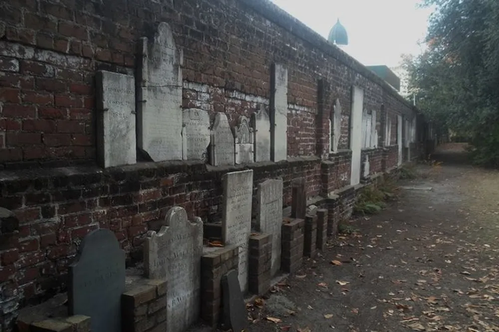 The image shows a series of weathered headstones leaning against a brick wall possibly indicating an old or historic cemetery