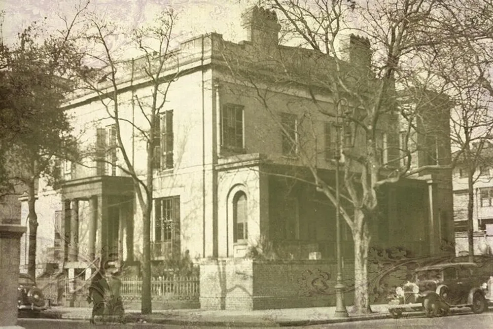 The image shows an old sepia-toned photograph of a two-story classical building with columns surrounded by a wrought iron fence bare trees and vintage automobiles invoking a sense of the early 20th century