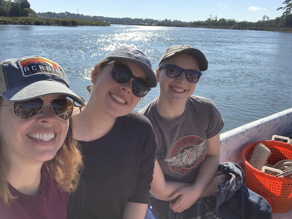 Three individuals are smiling for a selfie on a sunny day while seated on a boat with a river and greenery in the background