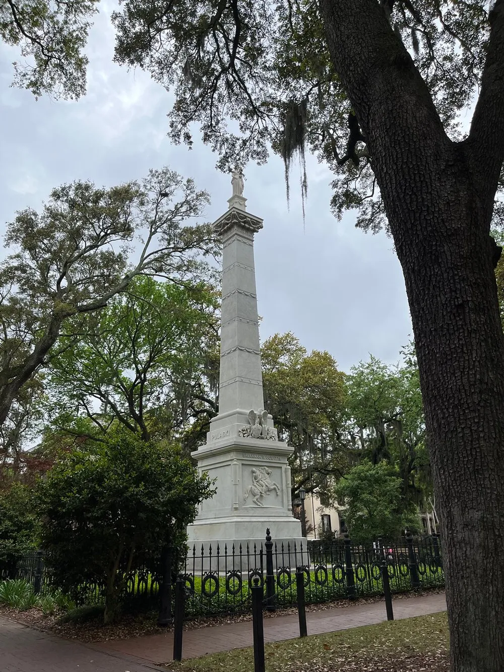 An impressive monument stands tall amidst a serene park setting with lush trees and Spanish moss enclosed by a decorative iron fence