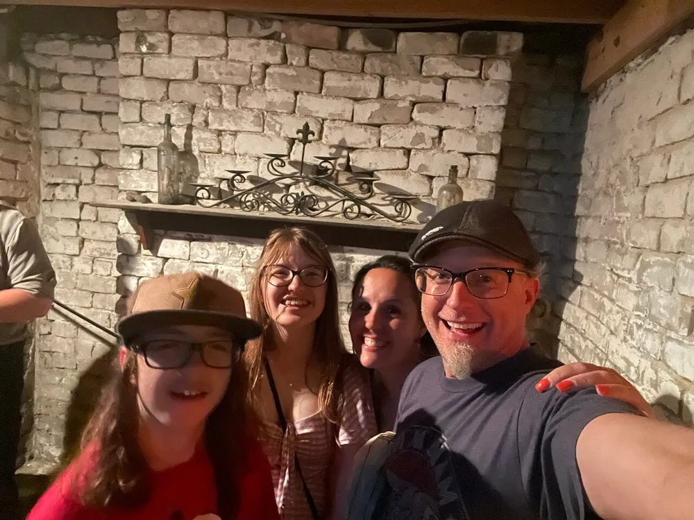 Four smiling people are taking a selfie together in front of a rustic stone wall with a decorative iron candelabra on a mantle